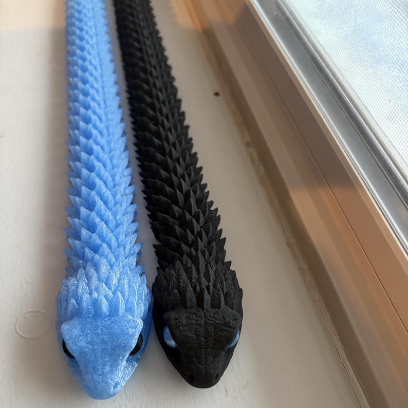 May include: Two 3D-printed articulated snake toys. One is light blue, the other is black. Both have a textured, scale-like pattern and small, blue eyes. The snakes are lying on a white surface.