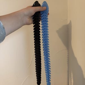 May include: Two articulated, 3D-printed tails are displayed. One is black, the other is light blue. Each tail is composed of numerous, leaf-like segments, creating a flexible, textured appearance. The tails are held against a neutral wall.