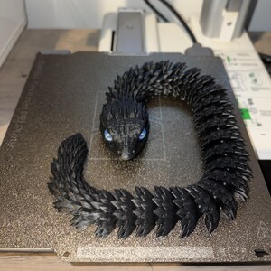 May include: A black articulated snake figurine with blue eyes. The snake is made of many small, textured segments, giving it a detailed, scaly appearance. The snake is coiled on a gray surface.
