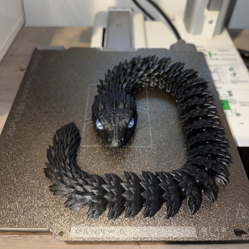 May include: A black articulated snake figurine with blue eyes. The snake is made of many small, textured segments, giving it a detailed, scaly appearance. The snake is coiled on a gray surface.