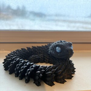 May include: A black articulated snake toy with a textured, spiky body and blue eyes. The toy is coiled and appears to be made of plastic. The snake is resting on a white surface near a window.