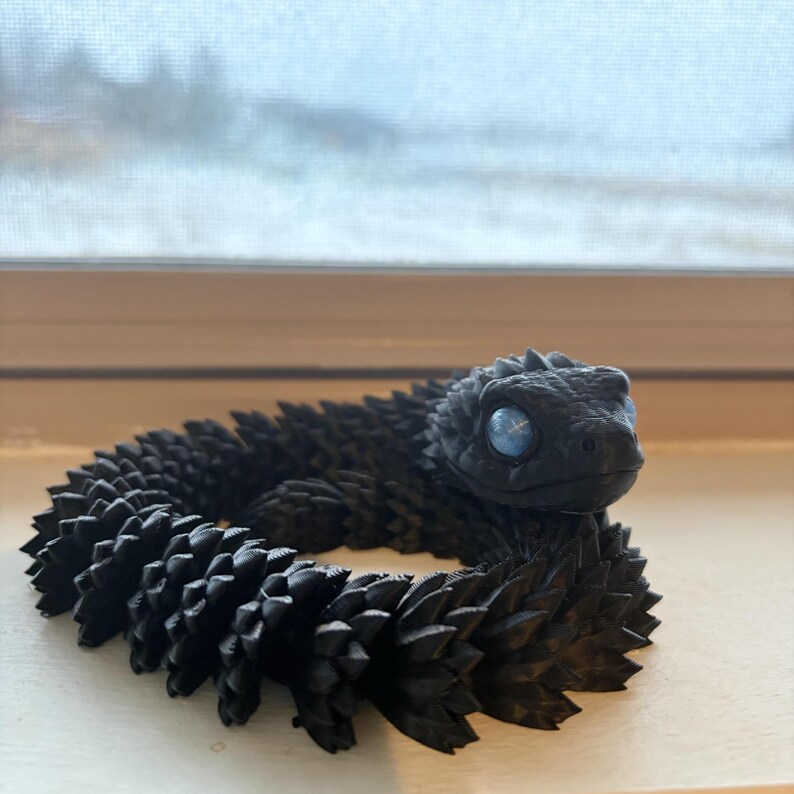 May include: A black articulated snake toy with a textured, spiky body and blue eyes. The toy is coiled and appears to be made of plastic. The snake is resting on a white surface near a window.