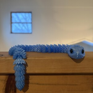 May include: A light blue, articulated snake toy with black eyes. The snake is resting on a wooden surface. The snake's body is segmented, with a textured, scale-like pattern. The tail has a unique, spiky design.