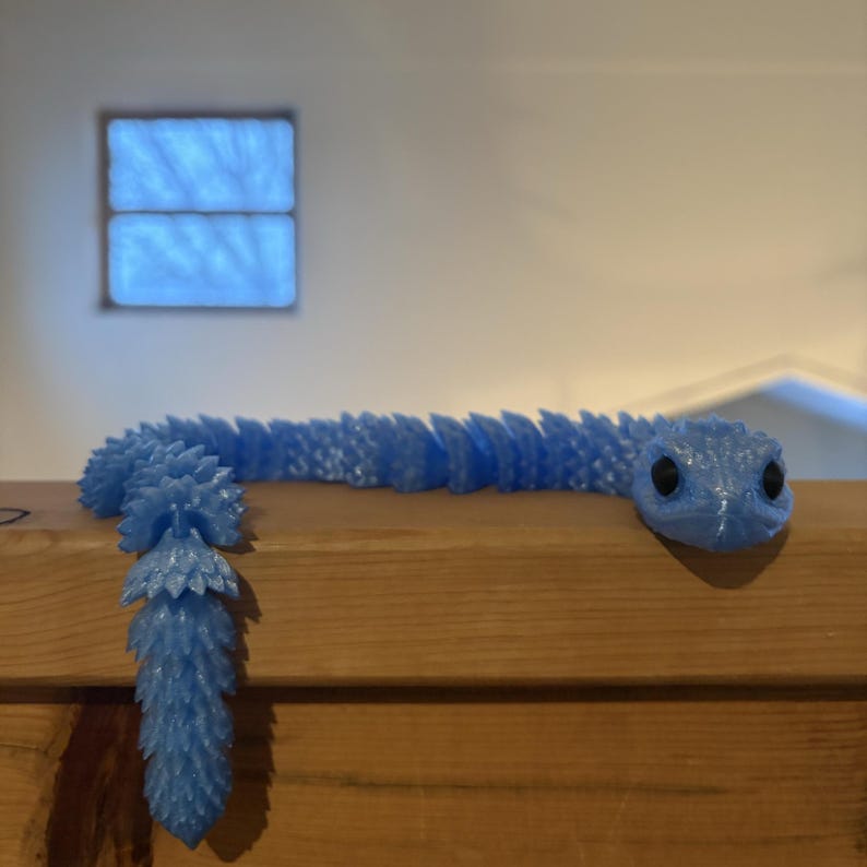 May include: A light blue, articulated snake toy with black eyes. The snake is resting on a wooden surface. The snake's body is segmented, with a textured, scale-like pattern. The tail has a unique, spiky design.