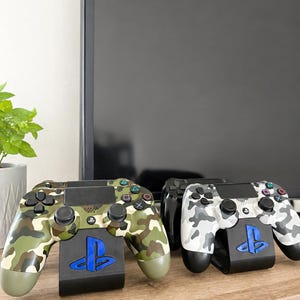 Op de afbeelding: Twee PlayStation 4-controllers met camouflagepatronen en oplaadstations. Eén controller heeft groen, bruin en beige camouflage, de andere grijs en wit. De stands zijn zwart met een blauw PlayStation-logo.