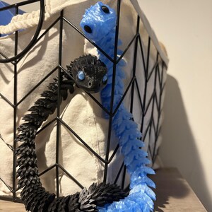 May include: Two articulated snake toys, one black and one light blue, are partially visible. The black snake is in the foreground, with the blue snake behind it. Both have detailed scales and are positioned near a black wire basket.