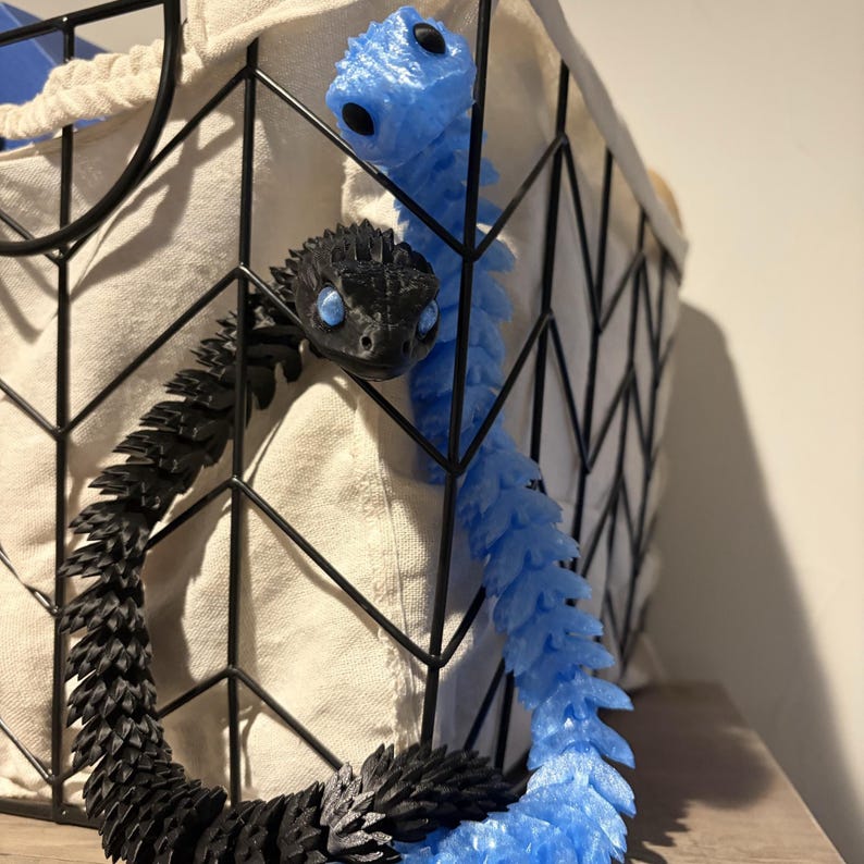 May include: Two articulated snake toys, one black and one light blue, are partially visible. The black snake is in the foreground, with the blue snake behind it. Both have detailed scales and are positioned near a black wire basket.