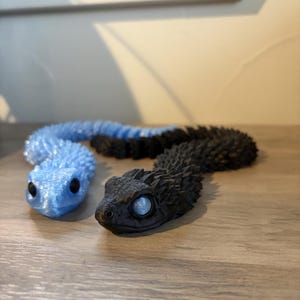 May include: Two articulated snake toys. One is light blue with black accents, the other is black with blue accents. Both have detailed scales and large, round eyes. The toys are lying on a wooden surface.