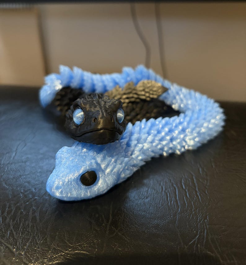 May include: Two 3D-printed toy snakes. One is black with silver eyes and a textured body. The other is light blue with a smooth body. The blue snake is coiled, and the black snake rests on top. The snakes are on a dark surface.