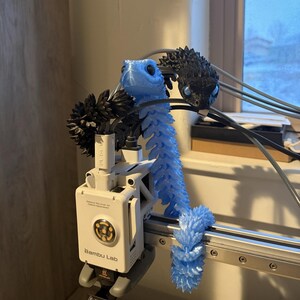 May include: A light blue articulated dragon toy with a spiky tail and a matching head. The dragon is 3D printed and is attached to a white Bambu Lab machine. A black dragon head is also visible.