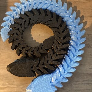 May include: A segmented, stylized snake sculpture in black and light blue. The body is composed of repeating leaf-like shapes, with the head and tail in a lighter blue. The sculpture is designed to be flexible and poseable.