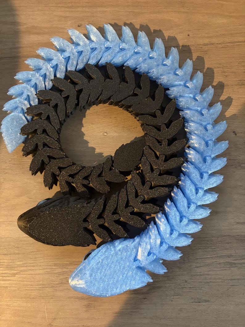 May include: A segmented, stylized snake sculpture in black and light blue. The body is composed of repeating leaf-like shapes, with the head and tail in a lighter blue. The sculpture is designed to be flexible and poseable.