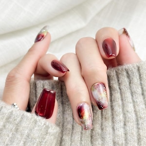 May include: Close-up of a hand with long, square-shaped nails. The nails are painted in a deep burgundy color, with some featuring a marbled design of gold, white, and red. The hand is resting on a gray sweater.