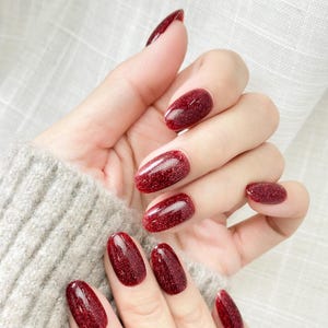 May include: Close-up of a hand with long, oval-shaped nails painted a deep red with glitter. The nails are glossy and reflect light, creating a sparkling effect. The hand is wearing a gray sweater.