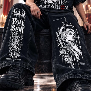 Op de afbeelding: Zwarte spijkerbroek van denim met witte grafische prints. De linkerpijp toont een kroon, een zwaard, rozen en de tekst PALE STAR en BREAK THE STRINGS. De rechterpijp bevat een illustratie van een persoon met een zwaard, rozen en de tekst CHOICE HUNGER ASCENT.