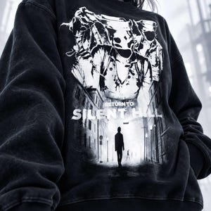 Może przedstawiać: Czarna bluza z okrągłym dekoltem i białym nadrukiem graficznym z napisem "RETURN TO SILENT HILL". Wzór przedstawia postać idącą zamgloną ulicą w stronę dużej pielęgniarki z wieloma oczami, która znajduje się w tle grafiki.