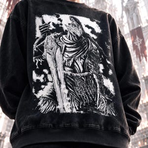 Könnte beinhalten: Ein schwarzes Acid-Wash-Sweatshirt mit einem großen, kontrastreichen weißen Grafikdruck auf der Vorderseite. Die Illustration zeigt einen Ritter in detaillierter Rüstung, der ein großes Schwert hält, umgeben von abstrakten, wolkenartigen Formen.