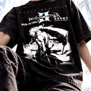 May include: A black acid-wash t-shirt featuring a white graphic print of a character with a sword from Devil May Cry 3. The design includes the text Devil May Cry 3, Japanese characters, and the name Chayamachi Suguro in a bold, high-contrast style.