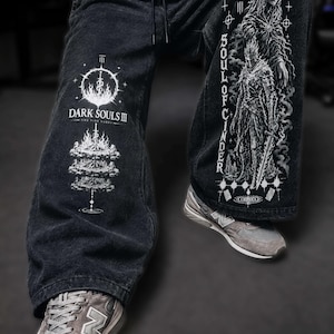 Könnte beinhalten: Schwarze Jeanshose mit weißen Grafikdrucken. Das rechte Bein zeigt das Dark Souls III Logo und eine Lagerfeuer-Illustration. Das linke Bein zeigt ein Soul of Cinder Charakterdesign mit dem vertikalen Schriftzug Soul of Cinder.