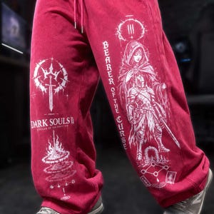 May include: Deep red sweatpants featuring white graphic prints. The left leg displays the Dark Souls II Scholar of the First Sin logo and a sword icon. The right leg features a hooded figure, a knight, and the text Bearer of the Curse.