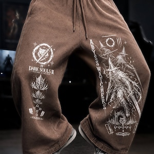 Könnte beinhalten: Braune Jogginghose mit weißen Grafikdrucken. Das linke Bein zeigt das Logo und die Symbole von Dark Souls III Watchers of the Abyss. Das rechte Bein zeigt eine Illustration der Abyss Watchers mit dem vertikalen Text Abyss Watchers.
