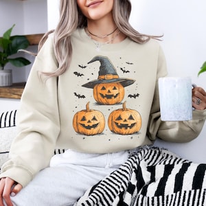 Op de afbeelding: Beige sweatshirt met een Halloween-ontwerp. Het ontwerp toont drie oranje pompoenen, waarvan één met een heksenhoed, met vleermuizen. De sweatshirt heeft een neutrale kleur, ideaal voor de herfst en Halloween.