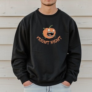 Può includere: Felpa girocollo nera con un design a forma di zucca dei cartoni animati. La zucca arancione ha una bocca aperta con denti. Il testo "FRIGHT NIGHT" è arcuato sotto in lettere arancioni.