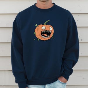 May include: Navy blue sweatshirt with a cartoon pumpkin design. The pumpkin is orange with a wide, toothy grin and green stem. The sweatshirt is a solid colour and has a crew neck.
