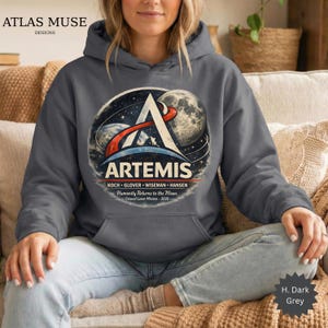May include: Dark grey hooded sweatshirt featuring the Artemis mission logo. The design includes a stylized 'A' with a space and moon graphic, and the text 'ARTEMIS' and 'Humanity Returns to the Moon'. The sweatshirt is a dark grey color.