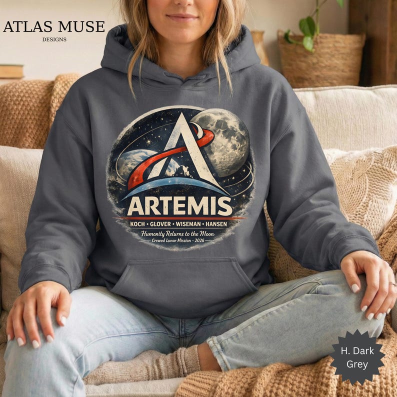 May include: Dark grey hooded sweatshirt featuring the Artemis mission logo. The design includes a stylized 'A' with a space and moon graphic, and the text 'ARTEMIS' and 'Humanity Returns to the Moon'. The sweatshirt is a dark grey color.