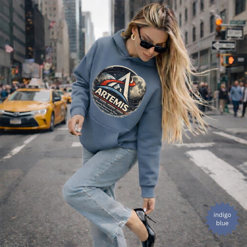 May include: Indigo blue hoodie with a large graphic featuring the Artemis logo, a stylized depiction of a rocket, Earth, and the moon. The hoodie has a kangaroo pocket and a drawstring hood. The text "ARTEMIS" is prominently displayed.
