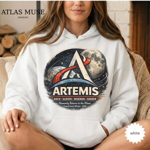 May include: White hoodie featuring the Artemis mission logo with a stylized A, planets, and the moon. The design includes the text "ARTEMIS" and the names of the crew members. The hoodie also has the text "Humanity Returns to the Moon" and "Crewed Lunar Mission - 2026".