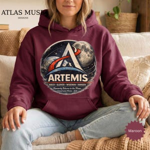 May include: Maroon hooded sweatshirt featuring the Artemis mission logo. The design includes the Artemis name, a stylized A, a depiction of the moon, and the text "Humanity Returns to the Moon." The sweatshirt also has a front pocket.
