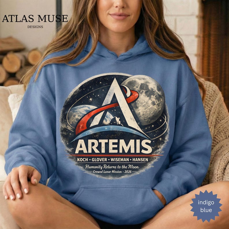 May include: Indigo blue hoodie featuring a large graphic with the word "ARTEMIS" and a space-themed design. The graphic includes a stylized "A", a moon, and the text "Humanity Returns to the Moon - Crewed Lunar Mission - 2026". The hoodie has a drawstring hood.