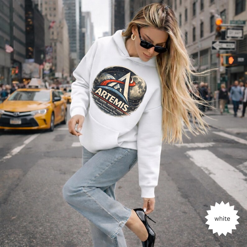 May include: White hooded sweatshirt featuring a graphic of the Artemis program, with a space-themed design including a moon and stars. The sweatshirt has a front pocket and long sleeves. The text "Artemis" is prominently displayed.