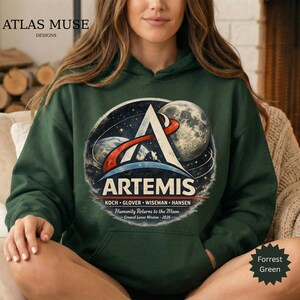 May include: A dark green hoodie featuring the Artemis logo with a space-themed design. The logo includes the word "ARTEMIS", the names of the crew, and the text "Humanity Returns to the Moon - Crewed Lunar Mission - 2026". The hoodie is a Forrest Green color.