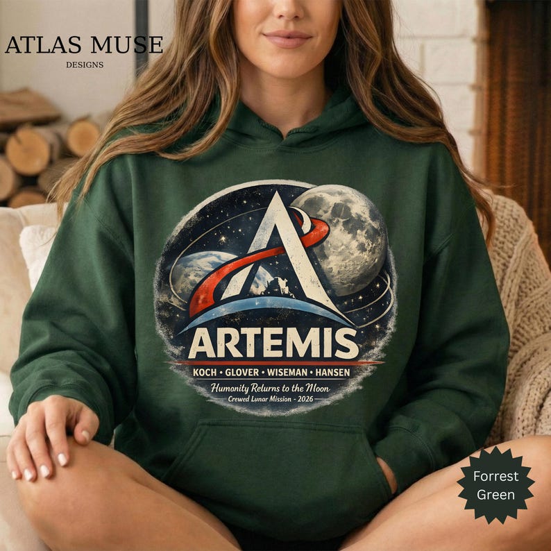 May include: A dark green hoodie featuring the Artemis logo with a space-themed design. The logo includes the word "ARTEMIS", the names of the crew, and the text "Humanity Returns to the Moon - Crewed Lunar Mission - 2026". The hoodie is a Forrest Green color.