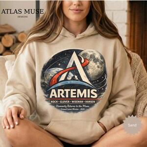 May include: Sand-colored hoodie featuring a large graphic of the Artemis mission. The design includes a stylized 'A' with a red and white arc, a depiction of the Earth and Moon, and the word 'ARTEMIS'. The names of the crew and mission details are also printed on the hoodie.