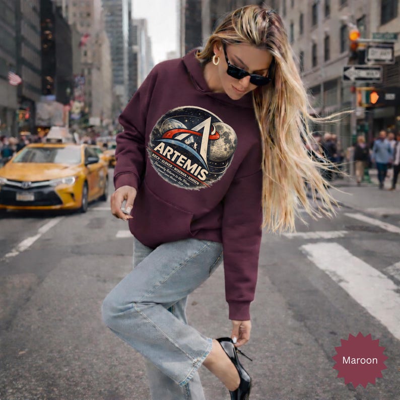 May include: Maroon hoodie with a large Artemis graphic on the front. The graphic features a stylized design with the word "Artemis" and other text. The hoodie has a hood and a front pocket. The background shows a city street.