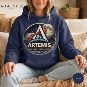 May include: Navy blue hooded sweatshirt featuring the Artemis mission logo. The design includes a stylized 'A' with a rocket trail, Earth, and the moon. Text on the sweatshirt reads "ARTEMIS" and "Humanity Returns to the Moon."