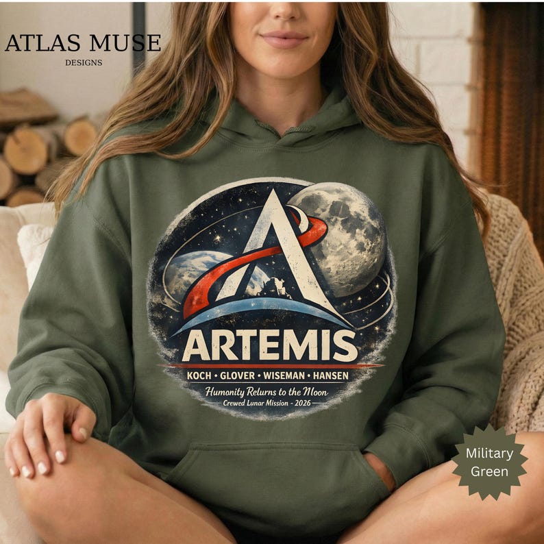 May include: Military green hoodie featuring the Artemis mission logo. The design includes a stylized 'A' with a red and white arc, a depiction of the Earth and Moon, and the text 'ARTEMIS'. Additional text includes the names of the crew and the mission details.
