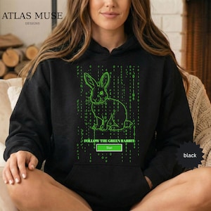 May include: Black hoodie featuring a neon green rabbit graphic with binary code and the text "FOLLOW THE GREEN RABBIT". The hoodie has a front pocket and a drawstring hood. The brand name "ATLAS MUSE DESIGNS" is printed at the top.