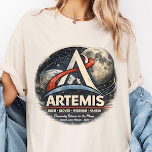 May include: Cream-colored t-shirt featuring a graphic of the Artemis mission. The design includes a large letter A, a moon, and the text "ARTEMIS" with the names of the crew members and the phrase "Humanity Returns to the Moon - 2026."