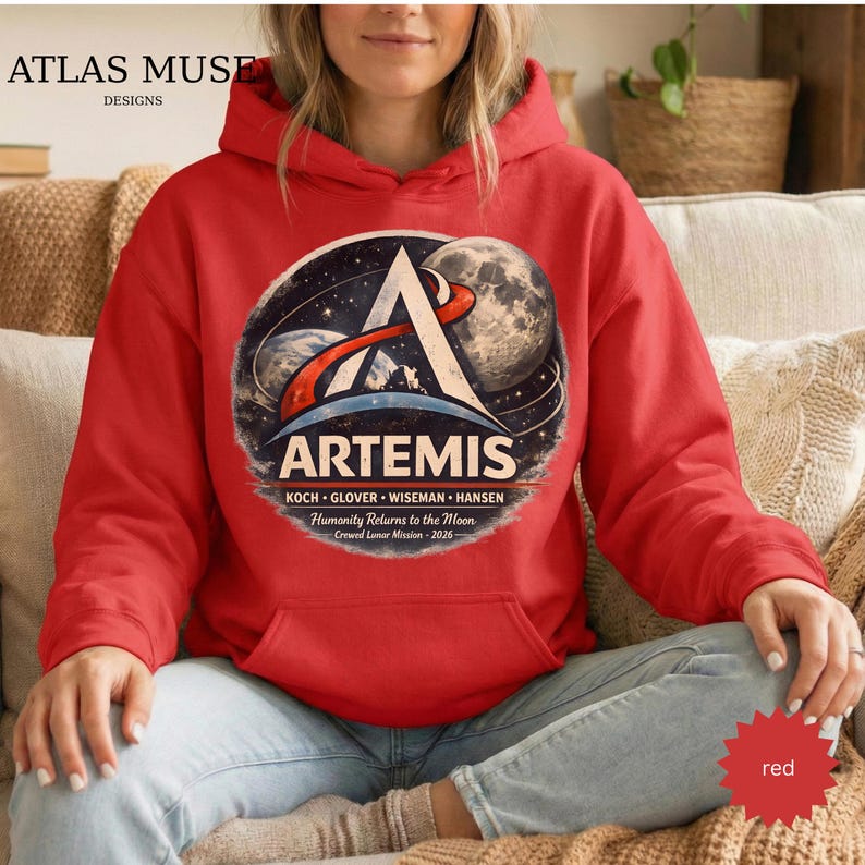 May include: A red hooded sweatshirt featuring a circular graphic with the word "ARTEMIS" and a space-themed design. The graphic includes the names of the crew and the text "Humanity Returns to the Moon Crewed Lunar Mission - 2026".