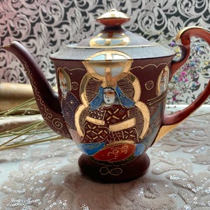 Puede incluir: Una tetera de cerámica marrón con tapa, boquilla y asa. La tetera está decorada con detalles dorados e ilustraciones de figuras. El asa y la boquilla son de un marrón más oscuro. La tetera está sobre una superficie blanca.