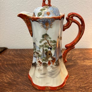 May include: A tall porcelain coffee pot featuring a white base with hand-painted scenes of figures in traditional attire among pine trees. The pot has a red and gold ornate handle, a matching lid with a decorative finial, and red trim.