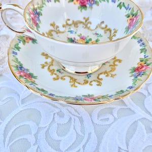 Peut inclure: Service à thé blanc avec des motifs floraux et dorés. La tasse et la soucoupe ont un bord doré et sont décorées de fleurs roses, bleues et jaunes. La tasse repose sur une nappe en dentelle blanche.