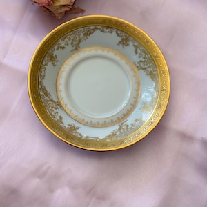 Puede incluir: Un platillo de taza de té vintage con un centro azul claro y un borde dorado. El platillo presenta intrincados diseños florales dorados y un círculo interior más pequeño. El borde dorado añade un toque de elegancia a esta delicada pieza.