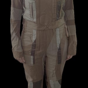 May include: A tan and brown jumpsuit featuring a high collar, long sleeves, and a waist belt. The garment includes quilted shoulder and knee patches, vertical chest pockets, and a multi-panel construction with varied fabric textures and tones.