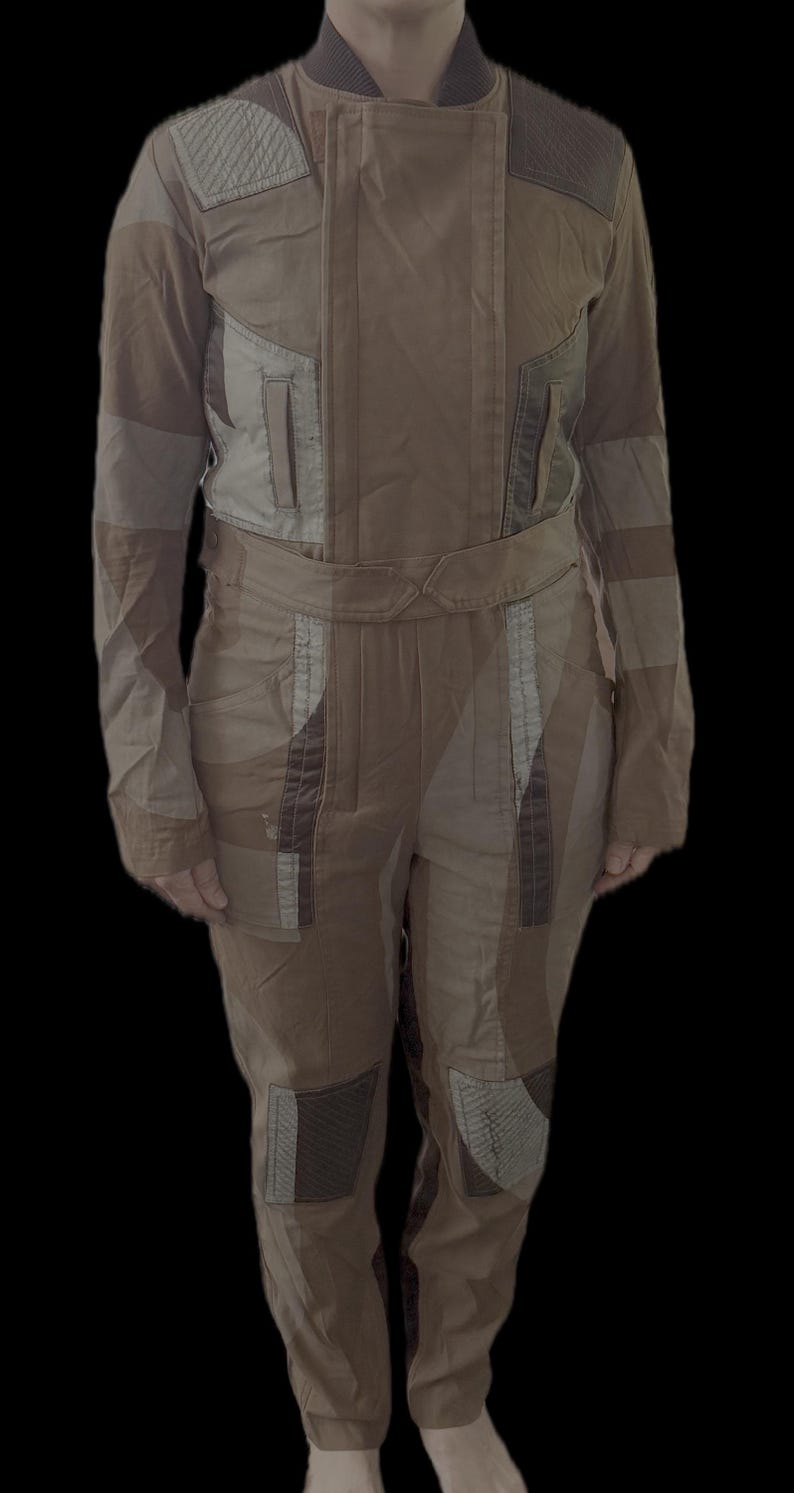 May include: A tan and brown jumpsuit featuring a high collar, long sleeves, and a waist belt. The garment includes quilted shoulder and knee patches, vertical chest pockets, and a multi-panel construction with varied fabric textures and tones.